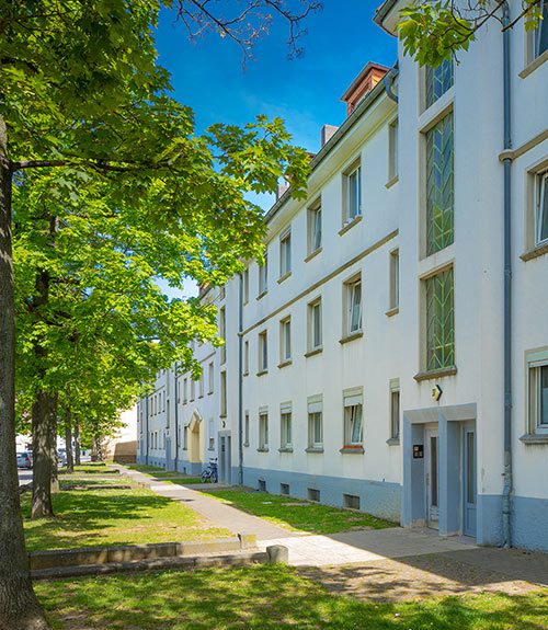 Das Foto zeigt einen Reihenbau an Mehrfamilienhäusern in Fulda. Grünfläche und Bäumen, sowie ein abgegrenzter Fußweg bieten den Häusern Privatsphäre von der Straße.